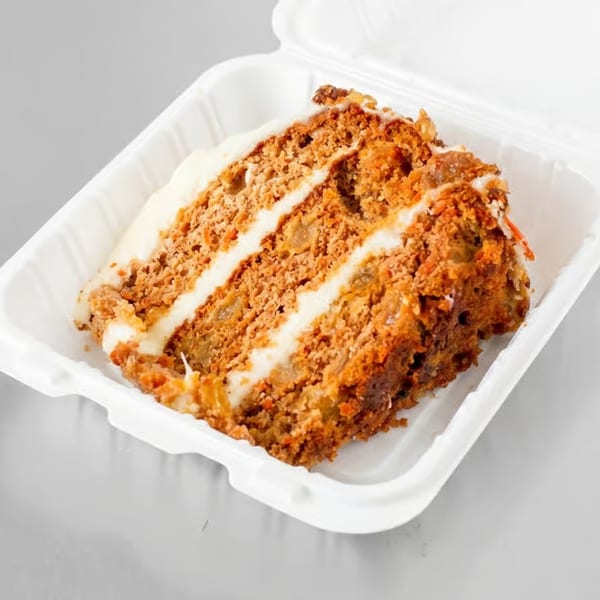 A slice of carrot cake with white frosting layers, in a white takeout container.