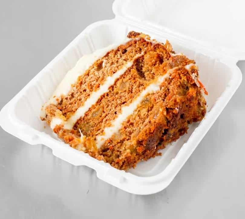 A slice of carrot cake with white frosting layers, in a white takeout container.